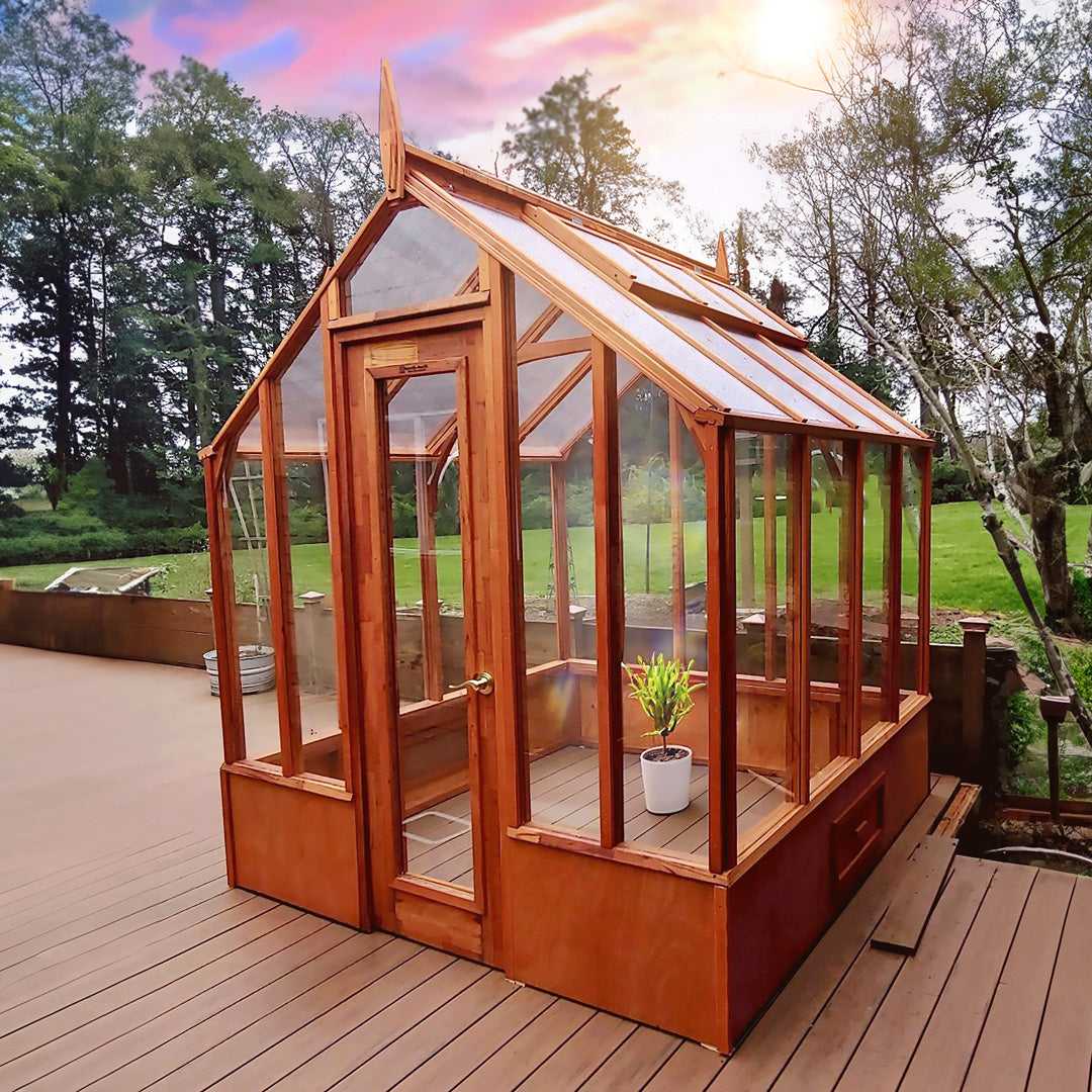 Wooden greenhouse with glass panels on a wooden deck, surrounded by trees and greenery.