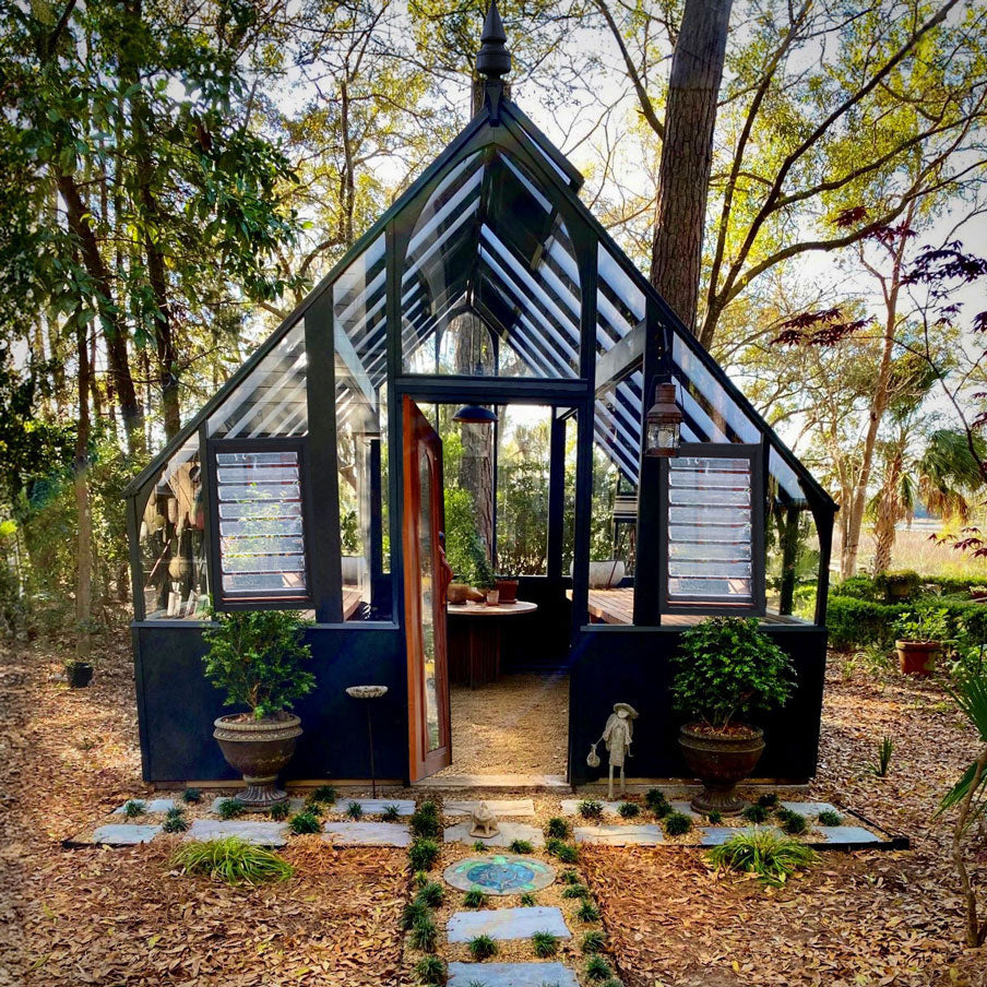 Glass greenhouse in a forest setting with potted plants and a stone path.