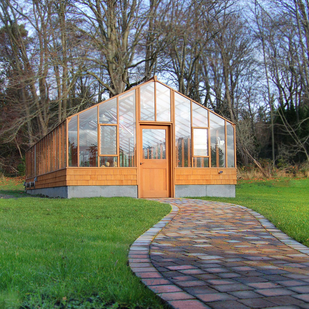 Modern greenhouse with wooden exterior and glass panels, surrounded by trees and a grassy area.