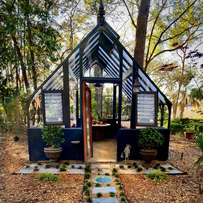 Glass greenhouse in a forest setting with potted plants and a stone path.